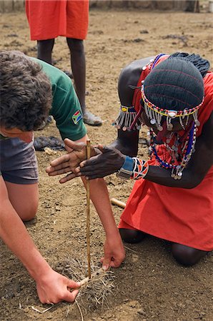 Kenya,Masai Mara National Reserve. A boy on a family safari assists a Maasai warrior as he demonstrates the art of making fire. Stock Photo - Rights-Managed, Code: 862-03366833