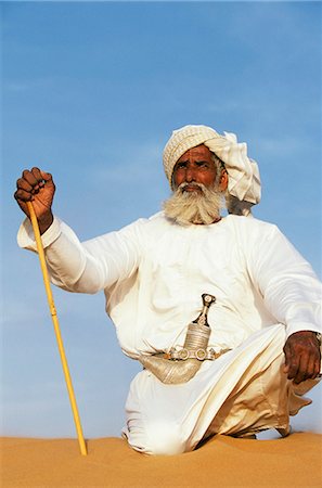A Bedouin man kneels on top of a sand dune in the desert . He wears the traditional Omani long white cloak or dish dash,a turban,a ceremonial curved dagger (khanjar) and holds a short camel stickOman 1John Warburton-Lee Foto de stock - Direito Controlado, Número: 862-03360154