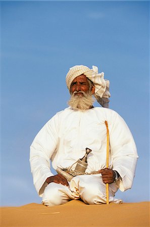 A Bedouin man kneels on top of a sand dune in the desert . He wears the traditional Omani long white cloak or dish dash,a turban,a ceremonial curved dagger (khanjar) and holds a short camel stickOman 2 JWLJohn Warburton-Lee Foto de stock - Con derechos protegidos, Código: 862-03360153