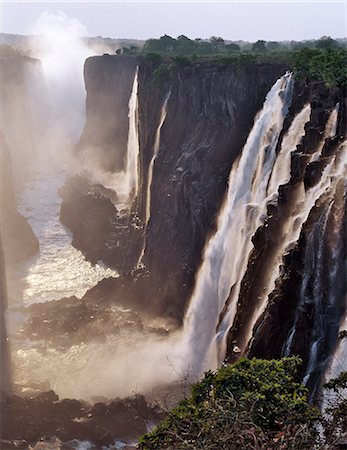 Late afternoon sunlight adds a glow to the magnificent Victoria Falls. The Falls are more than a mile wide and are one of the world's greatest natural wonders. The mighty Zambezi River drops over 300 feet in a thunderous roar with clouds of spray. Foto de stock - Con derechos protegidos, Código: 862-03438058