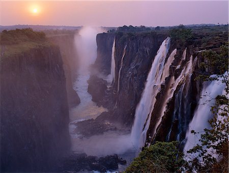 simsearch:862-03438042,k - Sunset over the magnificent Victoria Falls. The Falls are more than a mile wide and are one of the world's greatest natural wonders. The mighty Zambezi River drops over 300 feet in a thunderous roar with clouds of spray. Foto de stock - Direito Controlado, Número: 862-03438059