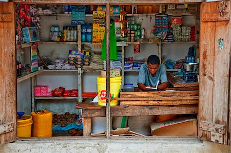 Tansania, Zanzibar. Ein Ladenbesitzer liest während des Wartens auf den Kunden an seinem kleinen Dorfladen in Nungwi. Stockbilder - Lizenzpflichtiges, Bildnummer: 862-03737293