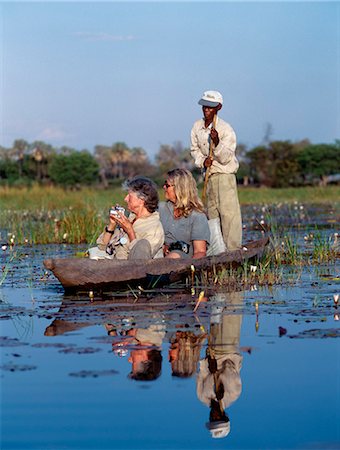 A boatman in his mokoro dug out canoe takes tourists game viewing along one of the myriad waterways of the Okavango delta.Fed by the Okavango River, which rises in Angola, the Okavango swamp covers an area of 6,500 square miles and is a haven for wildlife. Stock Photo - Rights-Managed, Code: 862-03820179