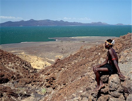 A young Turkana man looks out over Lake Turkana, often referred to as the Jade Sea due to the colour of its alkaline water. The barren, windswept country at this southeast corner of the lake is strewn with basalt lava boulders. Stock Photo - Rights-Managed, Code: 862-03820655