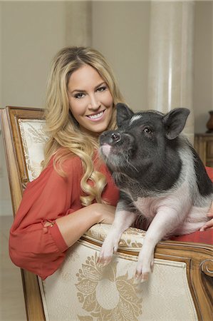 A miniature pot bellied pig sitting on his haunches on the lap of a blonde haired woman, sharing an antique armchair with thick cushions, in a large elegantly furnished mansion in Texas. Foto de stock - Con derechos protegidos, Código: 878-07442432